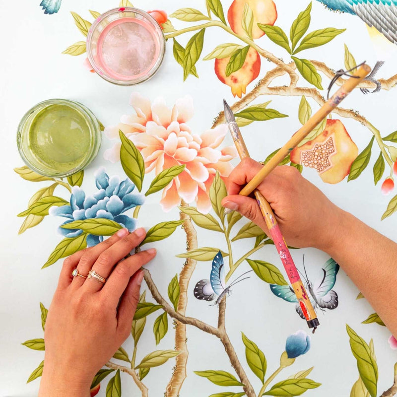 Up-close photo of Diane Hill's hands holding two chinoiserie paintbrushes over on of her ongoing chinoiserie paintings