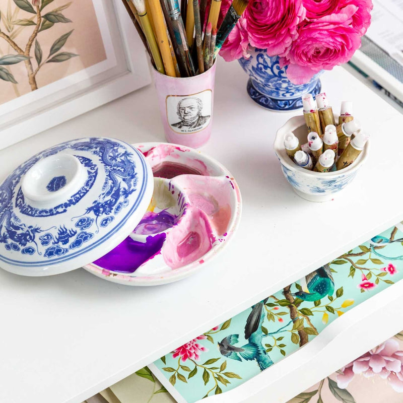 A paint palette, pot of paintbrushes and a bowl of paints next to flowers on top of a set of drawers that are open and contain Diane Hill's chinoiserie art prints