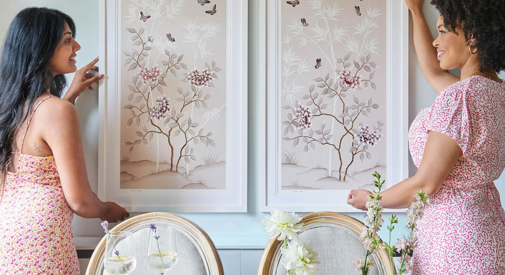 Diane Hill and friend hanging two matching dusty pink framed chinoiserie prints onto a wall behind a dining table and chairs
