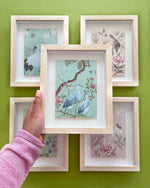 4 framed chinoiserie mini wall art prints in multi colours featuring botanical illustrations of birds, flowers, butterflies, and trees with hand holding one further framed mini art print featuring chinoiseriestyle herons, trees and flowers 