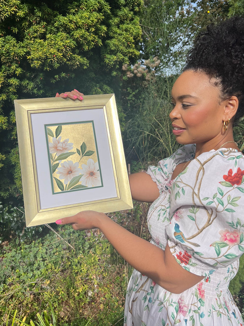 Diane Hill holding framed botanical gold leaf painting featuring 3 pink and white flowers 
