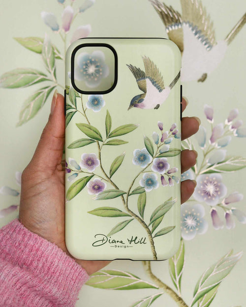 Hand holding luxury chinoiserie phone case featuring vintage inspired bird branches and flowers on a green background