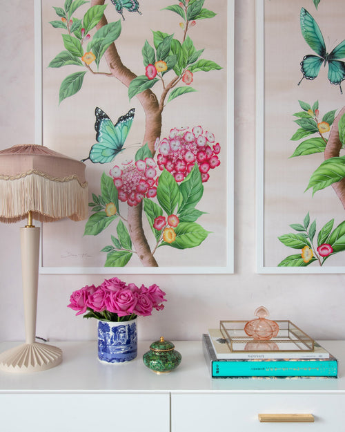 close up of pair of framed Chinoiserie style art prints featuring butterflies and pink flowers on a pastel pink background hung on wall