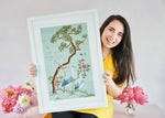 woman holding blue framed chinoiserie wall art print featuring antique inspired herons, flowers, and blossoms beneath a pine tree with butterflies