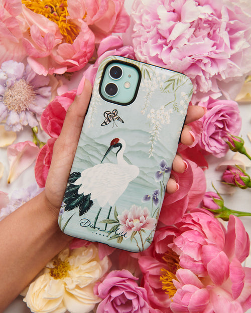 Hand holding luxury chinoiserie phone case featuring Japanese inspired crane, flowers, and wisteria on a blue mountain background