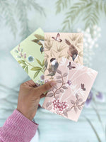 hand holding 3 chinoiserie mini wall art prints in multi colours featuring botanical illustrations of birds, flowers, butterflies, and trees