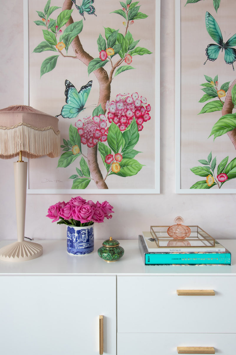 close up of pair of framed Chinoiserie style art prints featuring butterflies and pink flowers on a pastel pink background hung on wall