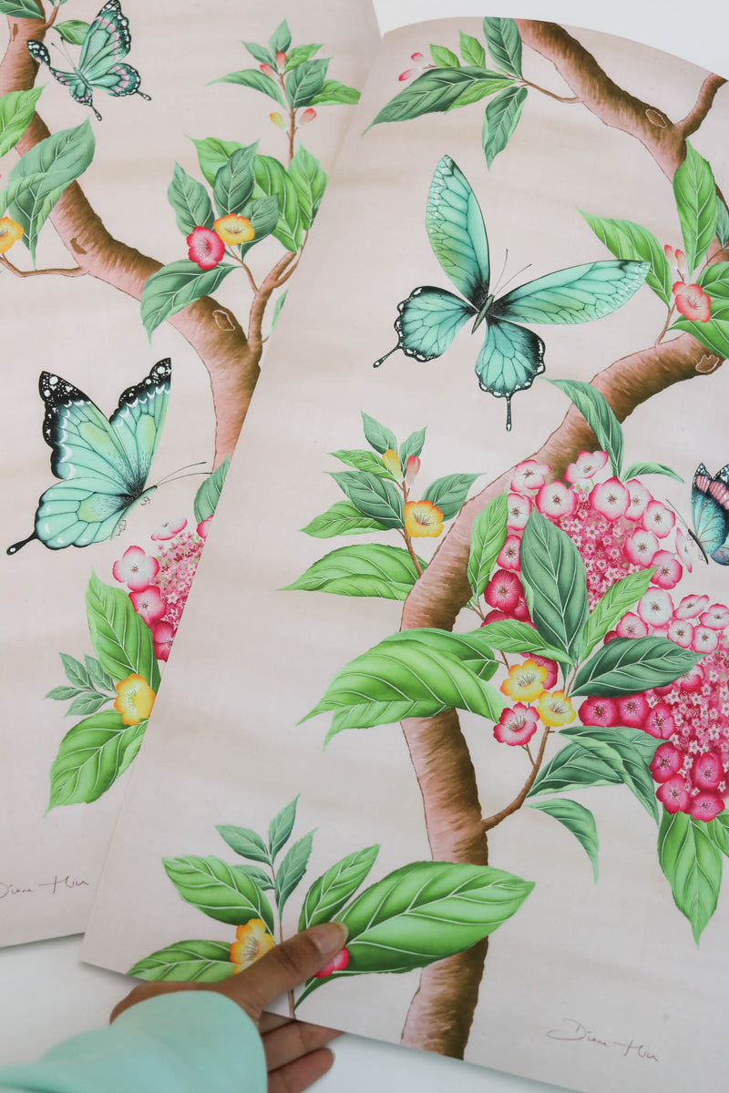 hand holding two Chinoiserie style art prints featuring butterflies and pink flowers on a pastel pink background
