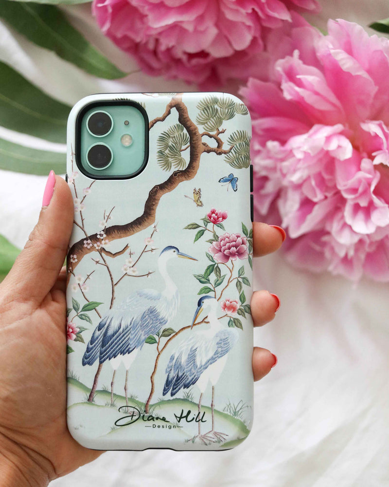 hand holding blue luxury chinoiserie phone case featuring antique inspired herons, flowers, and blossoms beneath a pine tree with butterflies