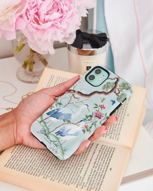 hand holding blue luxury chinoiserie phone case featuring antique inspired herons, flowers, and blossoms beneath a pine tree with butterflies