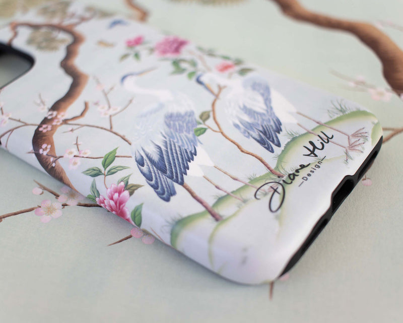 close up of blue luxury chinoiserie phone case featuring antique inspired herons, flowers, and blossoms beneath a pine tree with butterflies