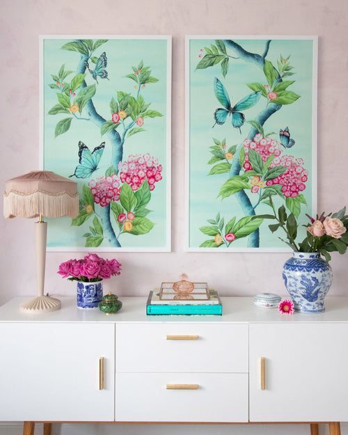 A pair of two framed Chinoiserie style art prints featuring butterflies and pink flowers on an aqua blue background hung on wall