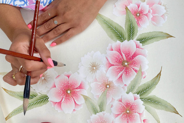 artist diane hill holding 2 Chinese paintbrushes next to her original floral painting on silk paper for her 'Rosa' chinoiserie wallpaper