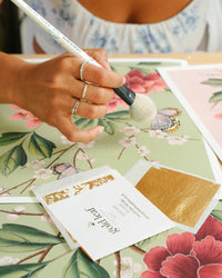 24 carat goldleaf being applied to pink chinoiserie art print featuring red flowers, cherry blossoms, and butterflies