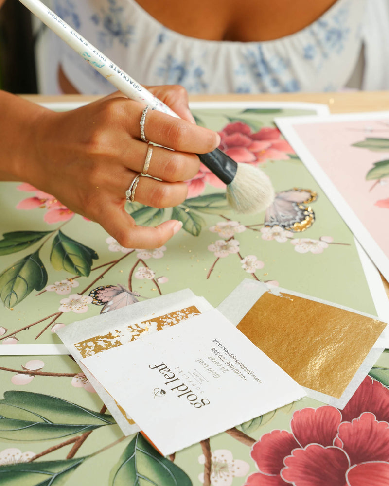 24 carat goldleaf being applied to pink chinoiserie art print featuring red flowers, cherry blossoms, and butterflies