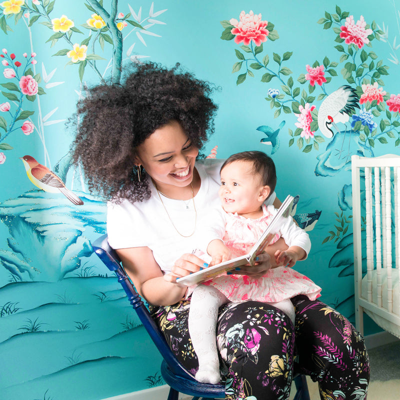 Chinoiserie artist Diane Hill sits with her daughter on her lap, in front of a bright and beautiful mural featuring birds, botanicals and plenty of colour, hand painted by Diane
