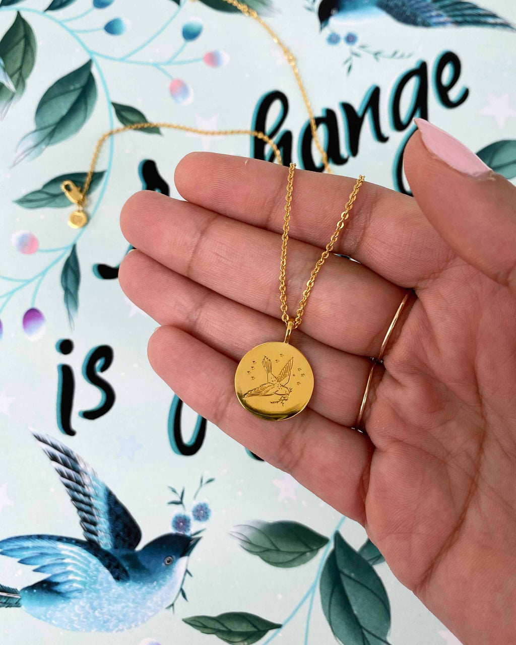 Diane Hill hill gently holds a gold pendant by Carrie Elizabeth Jewellery, featuring a little songbird in flight with a branch in its beak