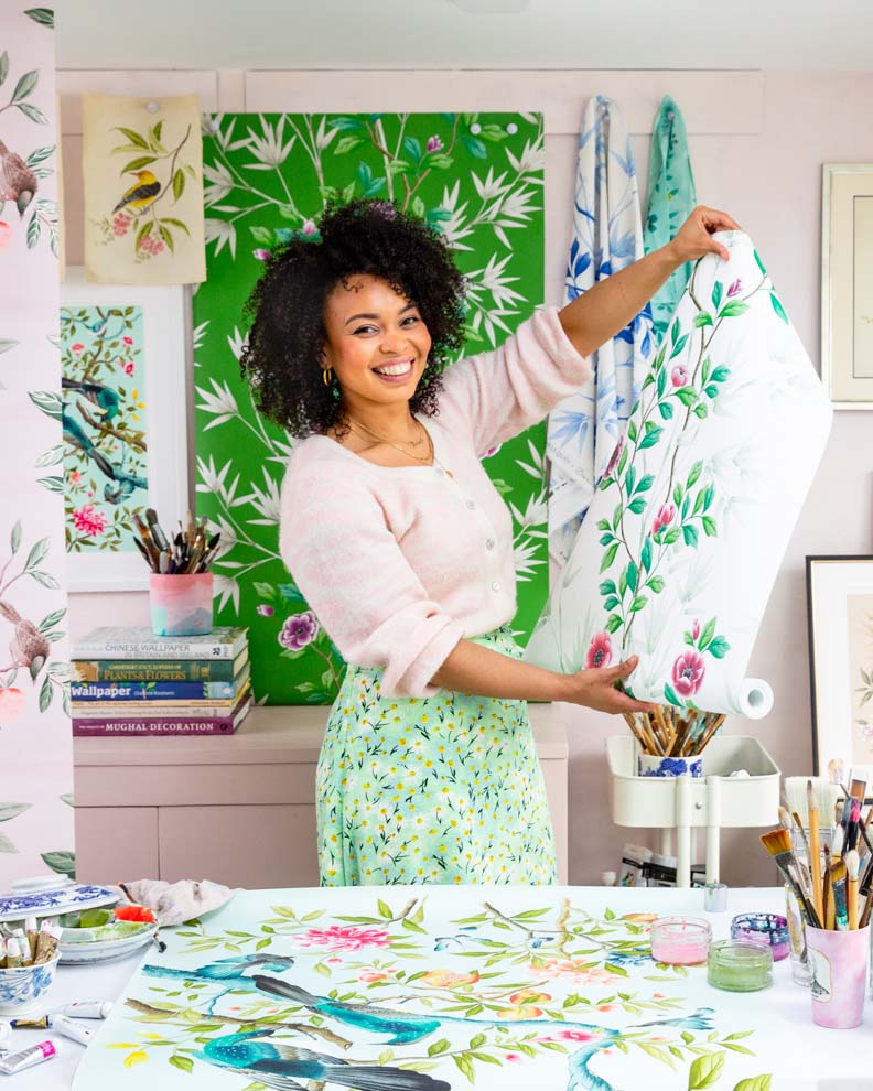 Chinoiserie artist Diane Hill holding up a roll of her chinoiserie wallpaper she designed for Harlequin in her art studio