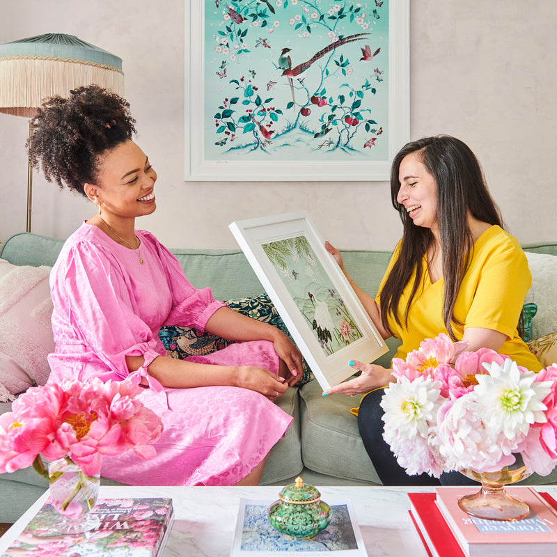 Chinoiserie artist Diane Hill sits on a sofa with a friend, holding a framed chinoiserie art print