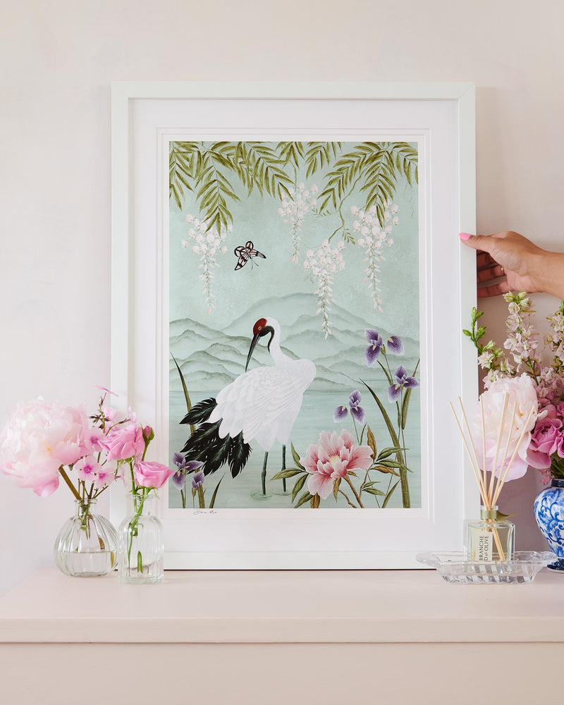 framed chinoiserie wall art print featuring Japanese inspired crane, flowers, and wisteria on a blue mountain background on bedside table