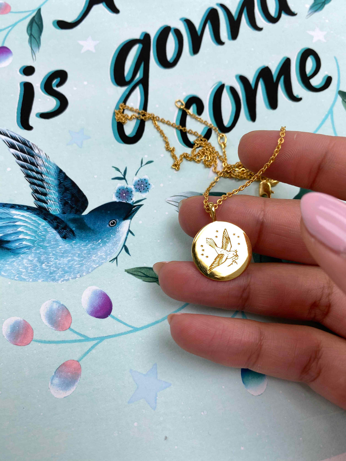 Diane Hill hill gently holds a gold pendant by Carrie Elizabeth Jewellery, featuring a little songbird in flight with a branch in its beak