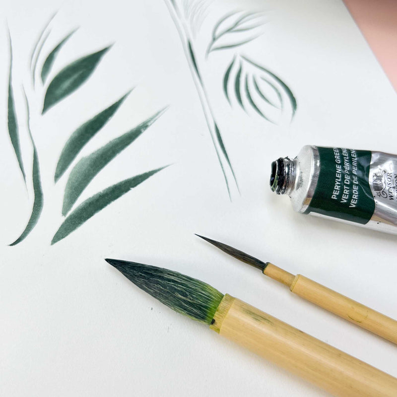 2 chinoiserie paintbrushes laying on white silk paper that has botanical shaped brush strokes painted onto it next to a tube of green gouache paint