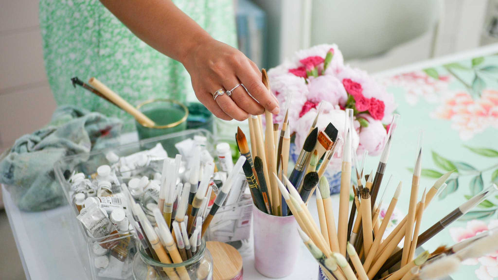 Chinoiserie paintbrushes in pots surrounded by paints and artwork in Diane Hill's art studio