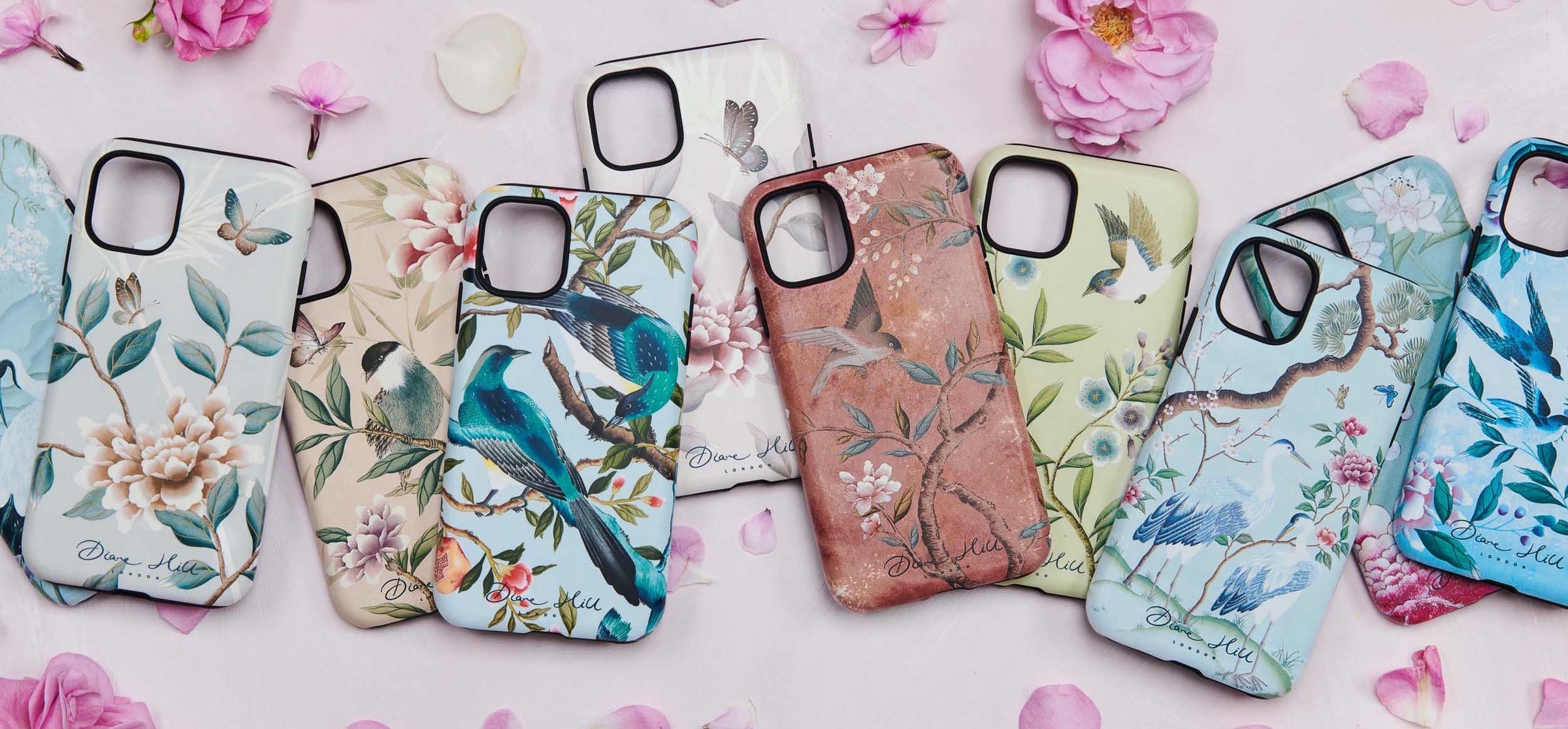 Various luxury phone cases featuring Diane Hill's chinoiserie designs spread across a pink background with pink flower petals around them