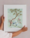 blue framed chinoiserie wall art print featuring antique inspired herons, flowers, and blossoms beneath a pine tree with butterflies hung on wall