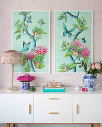 A pair of framed Chinoiserie style art prints by Diane Hill featuring large scale butterflies and pink flowers on an aqua blue background. Framed as a pair in a living room and hung over a console table 
