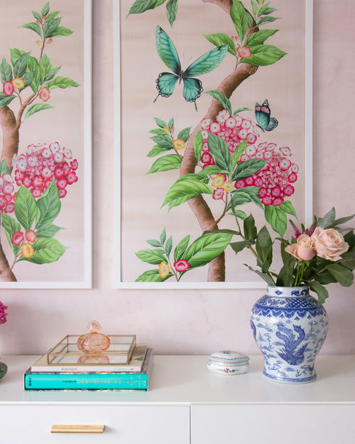 A pair of pink Chinoiserie art prints sit side by side over a modern sideboard. This pair of Chinoiserie panels are framed in a simple white frame. They feature pink hydrangea flowers, green leaves and large blue butterflies.
