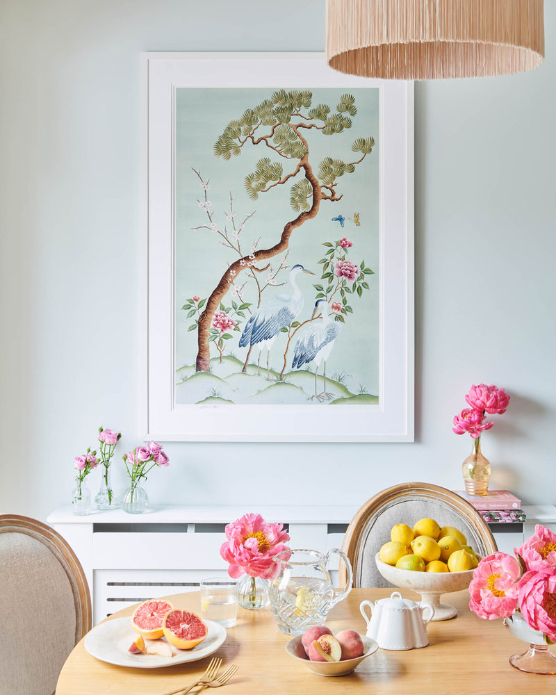 Susan Chinoiserie Art Print, framed in a classic white frame. Featuring herons, butterflies, blossom and peonies, this modern chinoiserie print by Diane Hill is inspired by a beautiful antique painting found in The National Trust’s Saltram House. The large Susan art print sits on a wall in a dining room with the dining table decorated with fresh lemons and pink peonies in vases.