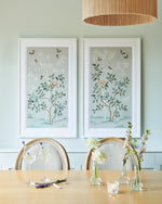 The Mariya art print in pebble blue, in a classic white frame. A celebration of butterflies, bamboo and botanicals, Mariya is a beautiful modern chinoiserie art print which was originally hand-painted onto pure silk, featuring delicate leaves, peonies in tones of blue and bronze, and an abundance of butterflies. The print is framed above a dining table and flowers