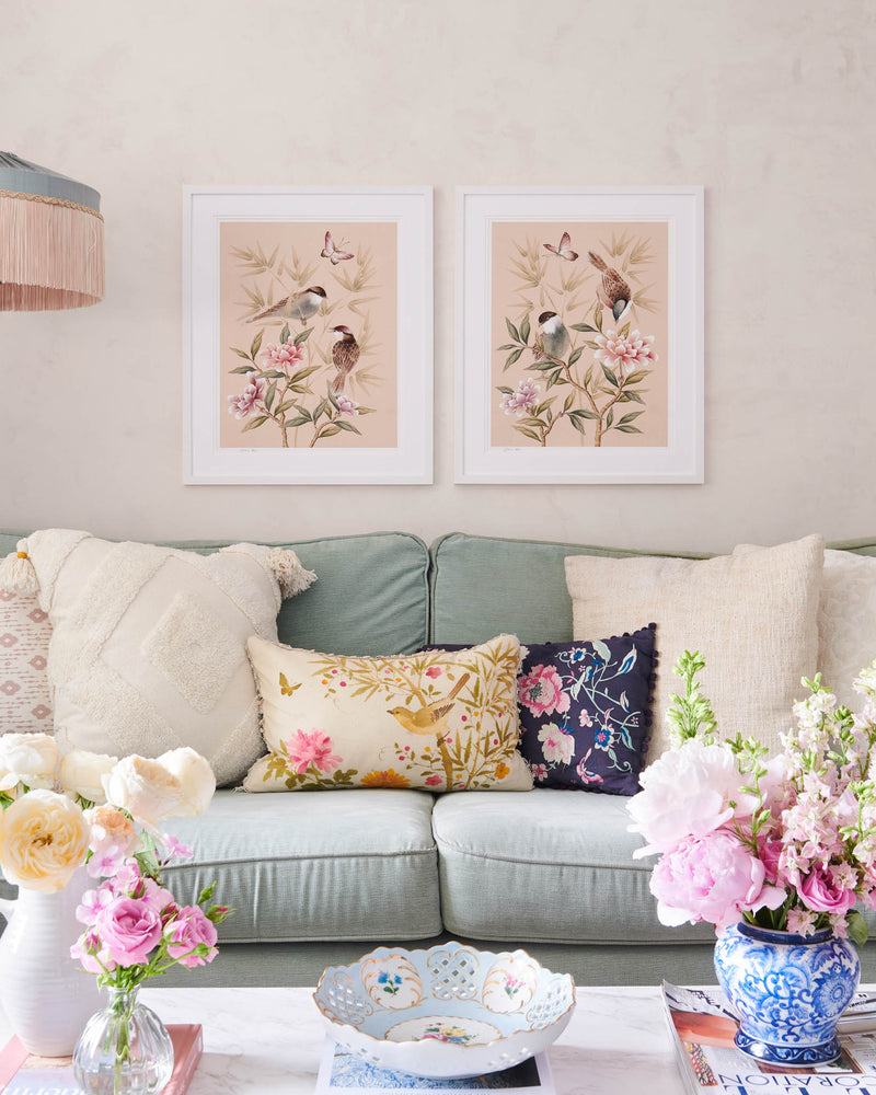 Rosie and Bonnie Chinoiserie Art Print, framed in a classic white frame against a peachy pink wall. The Rosie and Bonnie art prints by Diane Hill are beautifully restful, a calming chinoiserie style art print featuring soft colours which are hung side by side above a sofa in a living room. The room is decorated with soft floral cushions and fresh flowers sit on the coffee table in front.