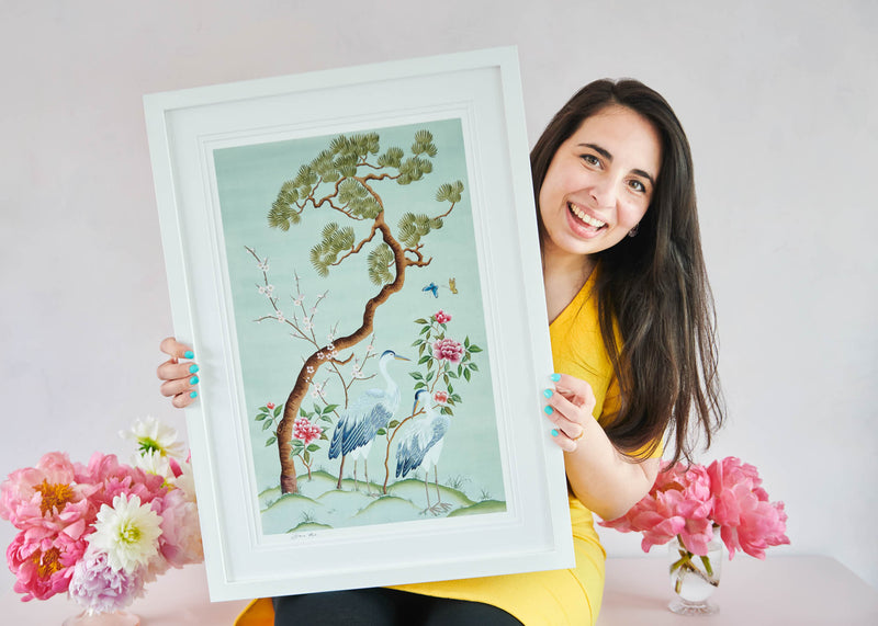 Diane Hill's Susan Chinoiserie art print in tones of blue , green and accents of pink, framed in a classic white frame. The art print is being held by a smiling woman in a pink room surrounded by beautiful fresh flowers.