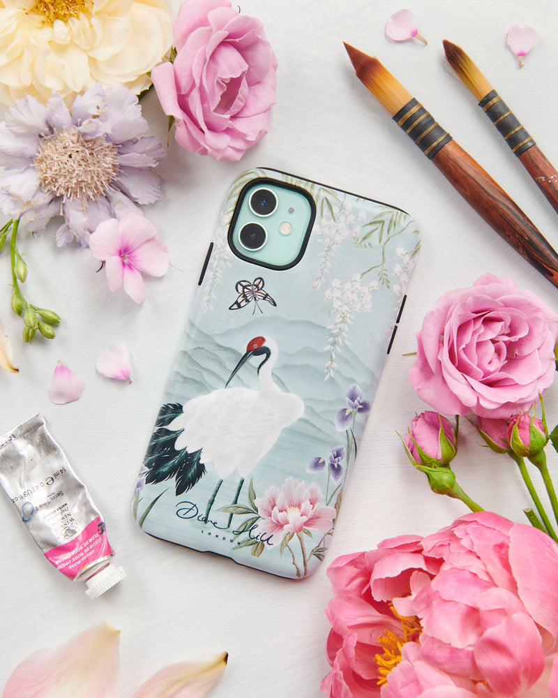 The Elina phone case by Diane Hill. Elina features a graceful white bird standing in front of a pale blue mountainscape, with butterflies and blossom overhead, and peonies and iris in the foreground. A styled flatlay set with peonies and paintbrushes