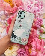 The Elina phone case by Diane Hill, with an abundance of peonies in the background. Elina features a graceful white bird standing in front of a pale blue mountainscape, with butterflies and blossom overhead, and peonies and iris in the foreground.