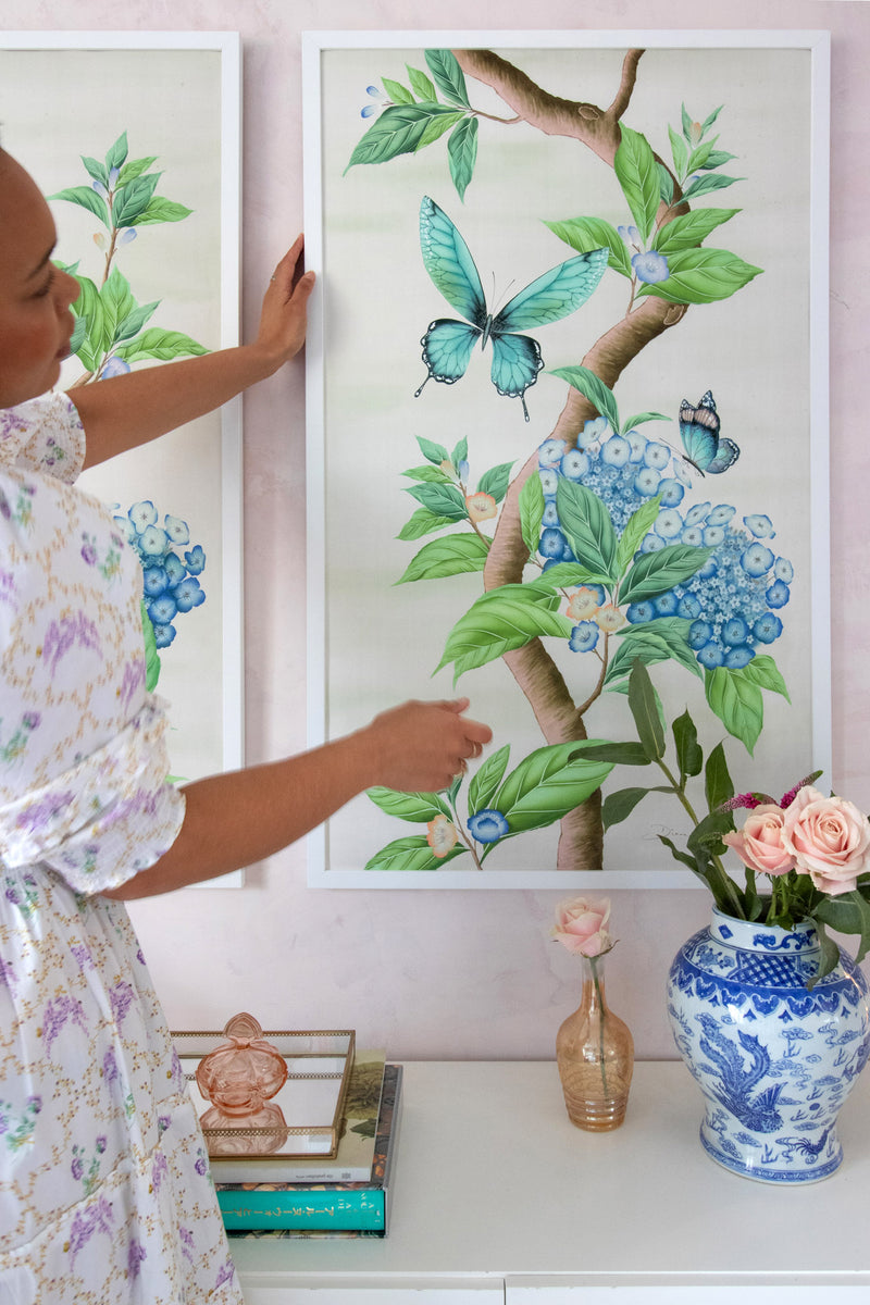 Diane Hill hanging a framed Chinoiserie art print on the wall above a styled console table. The design features botanical green leaves, delicate blue hydrangeas and large butterflies.