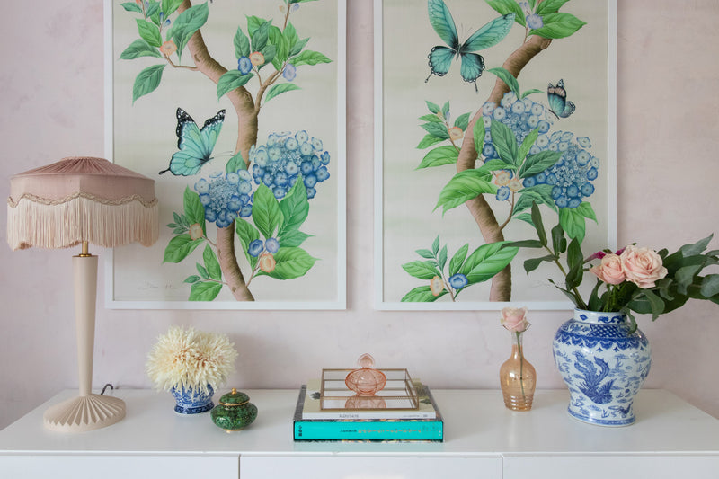 A pair of framed Chinoiserie art prints by Diane Hill in ivory neutral tones, featuring botanical green leaves, blue hydrangea flowers and fluttering butterflies. The two Chinoiserie style panels sit above a modern sideboard