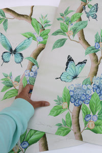 hand holding Chinoiserie style art prints featuring butterflies and blue flowers on an ivory cream background