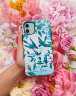 A hand holds the Julia phone case by Diane Hill, with an abundance of peonies in the background. The Julia design by Diane Hill is a bright and joyful modern chinoiserie design, featuring two swooping songbirds set against a turquoise background, framed by leaves and flowers