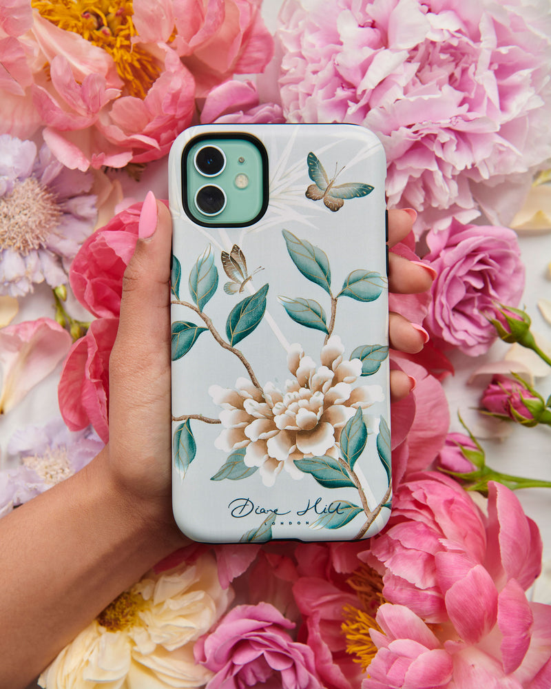 A hand holds the Lilly phone case by Diane Hill, with an abundance of peonies in the background. A celebration of butterflies, bamboo and botanicals, Lilly is a beautiful modern chinoiserie design which was originally hand-painted onto pure silk, featuring delicate leaves and an abundance of butterflies