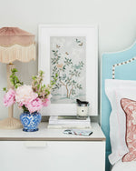 The Mariya art print in pebble blue, in a classic white frame. A celebration of butterflies, bamboo and botanicals, Mariya is a beautiful modern chinoiserie art print which was originally hand-painted onto pure silk, featuring delicate leaves, peonies in tones of blue and bronze, and an abundance of butterflies. The print is framed above a bedside table set with candles and flowers
