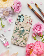The Rosie phone case by Diane Hill, featuring a pretty songbird perched on a branch with butterflies overhead and an abundance of blooming peonies in the background. A styled flatlay set with peonies and paintbrushes
