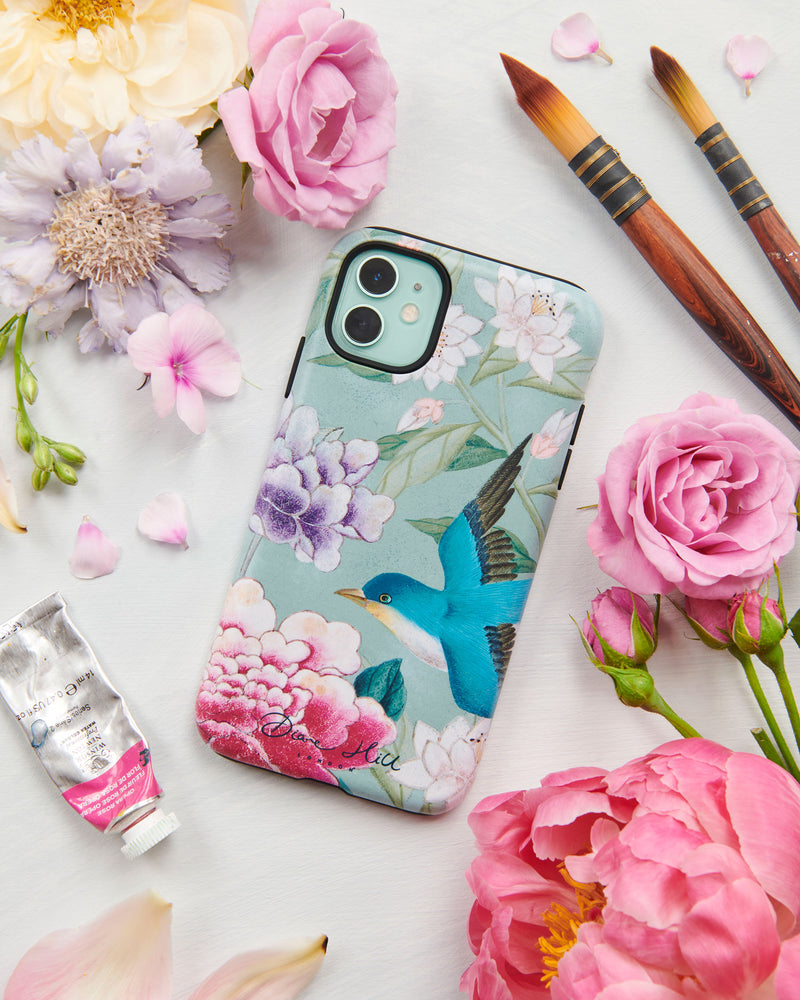 The Shona phone case by Diane Hill with an abundance of peonies in the background. Shona is a brilliantly colourful and elegant design with a subtle antique feel, featuring a joyful blue bird, water lilies, peonies and leaves. A styled flatlay set with peonies and paintbrushes