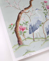 close up of blue chinoiserie wall art print featuring antique inspired herons, flowers, and blossoms beneath a pine tree with butterflies